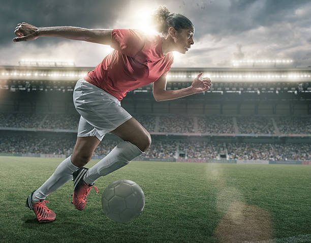 Female Soccer Superstar A mid action image of a female soccer player of African descent running and dribbling a soccer ball on an outdoor football pitch. The action takes place in a generic floodlit stadium full of spectators under a storm evening sky at sunset. The footballer is wearing a generic unbranded red and white soccer kit.  soccer striker stock pictures, royalty-free photos & images