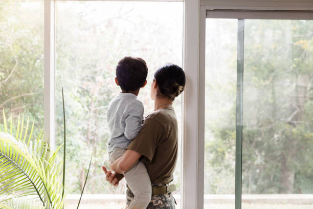 Female soldier looks through window with son Rear view of a female soldier looks through window while holding her preschool age son. veterans returning home stock pictures, royalty-free photos & images