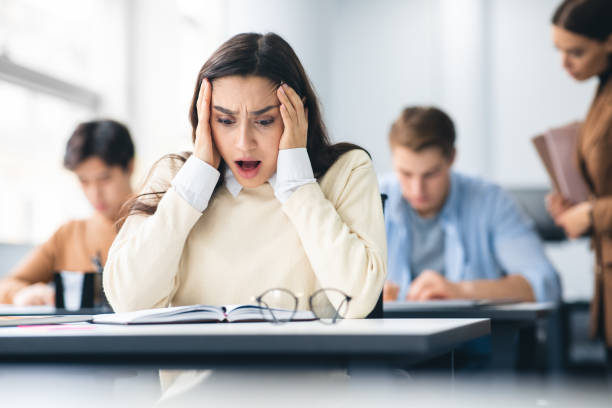 Female student grabbing head sitting at desk in class Portrait of shocked female student sitting at desk in classroom, grabbing her head looking at exam questions or results. Worried youth unprepared for test, thinking about deadline or low score hardest exams stock pictures, royalty-free photos & images