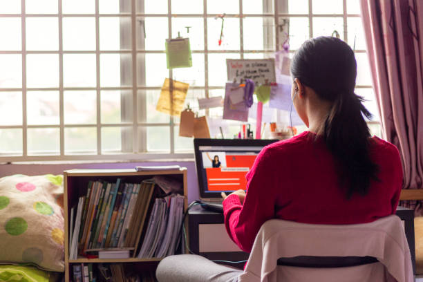 Gadis remaja Asia mengenakan earphone duduk di depan lapttop, menghadiri kelas video online dengan guru di kamar tidurnya