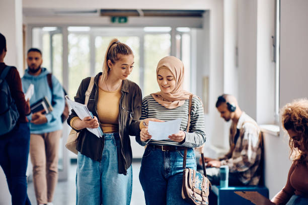 Female students going through lecture notes while walking through the hallway. Happy Muslim student and her friend reading their exam results while walking through university hallway.  student life stock pictures, royalty-free photos & images