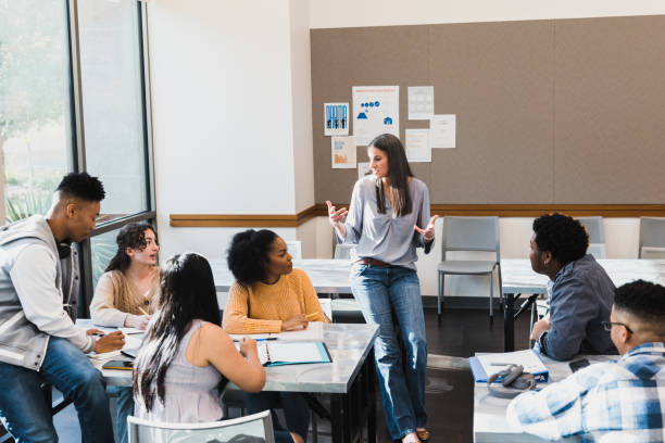 Female teacher leans against table and leads group discussion The mid adult female teacher leaning against the table leads an informal group discussion with the diverse high school students.  student life stock pictures, royalty-free photos & images