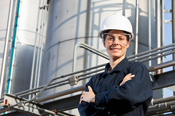 Female worker at an industrial plant A low angle view of a confident woman working at a chemical manufacturing plant, standing outdoors with arms crossed in front of industrial storage tanks.  She is wearing a white hardhat, safety goggles and dark blue coveralls.  She is in the minority as a female engineer working in a factory. Refinery stock pictures, royalty-free photos & images