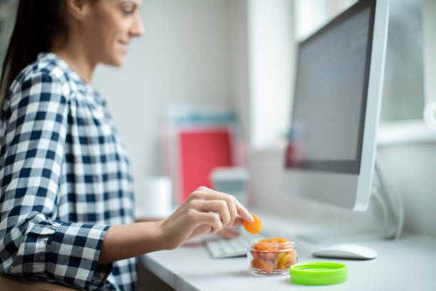 Female Worker In Office Having Healthy Snack Of Dried Apricots At Desk Female Worker In Office Having Healthy Snack Of Dried Apricots At Desk snack stock pictures, royalty-free photos & images