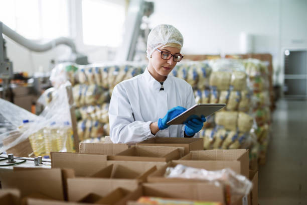 female worker using tablet for checking boxes while standing in food factory. - food imagens e fotografias de stock