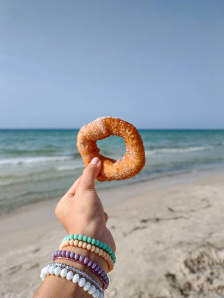 Femele hand holding doughnut on the beach. Female left hand holding donut on the beach. Sunny day. Hand with bracelets. Beach in background. tunisia woman stock pictures, royalty-free photos & images