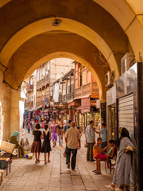 Fes Medina, Morocco Fes Medina, Morocco - July 12, 2010: People walk through an arched gateway in the historical medina or "Fas el Bali" which is a UNESCO world heritage site. People can be seen by the side of the street and some people near shops in the background. Fes is the former capital of Morocco and one of the countries four imperial cities. medina district stock pictures, royalty-free photos & images