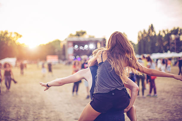 Festival vibes A young couple embracing at an outdoors music festival. Man carrying girlfriend. Stage in background. Summertime, evening. Caucasian ethnicity. View from behind. music festival stock pictures, royalty-free photos & images