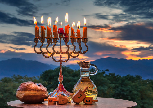 Festive donut, menorah, burning  candles and jar with olive oil - traditional symbols of Hanukkah Holiday. English translation for Hebrew letters on four sides of dreidel means - Great Miracle Happened There or Here. Blurred background with morning clouds, parks and mountains