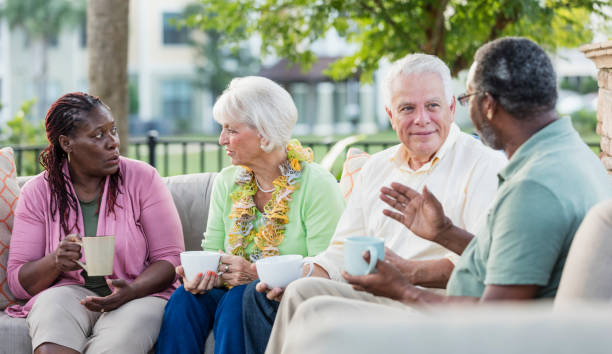 A multi-ethnic group of mature and senior friends hanging out together, sitting outdoors on a patio conversing, drinking coffee. The African-American couple are in their 50s. The Caucasian couple are in their 70s. The women are sitting next to each other, talking, and the men are chatting with each other.