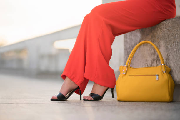 Fashion portrait of an unrecognizable young woman sitting at the balcony in red trousers and high heels, with a yellow bag next to her; cut out.