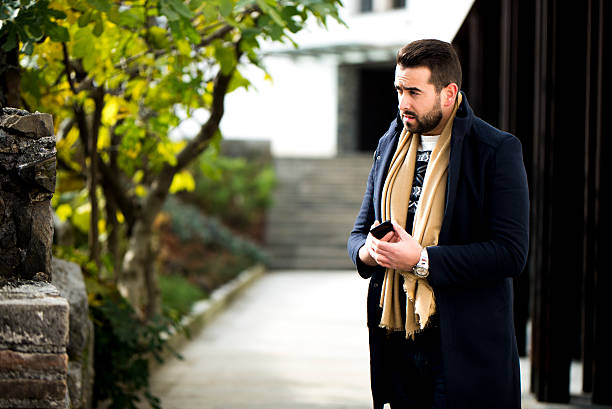Young man with a bierd standing outdoors, wearing a black coat and a beige scarf and holding a smart phone in his hands while looking away.
