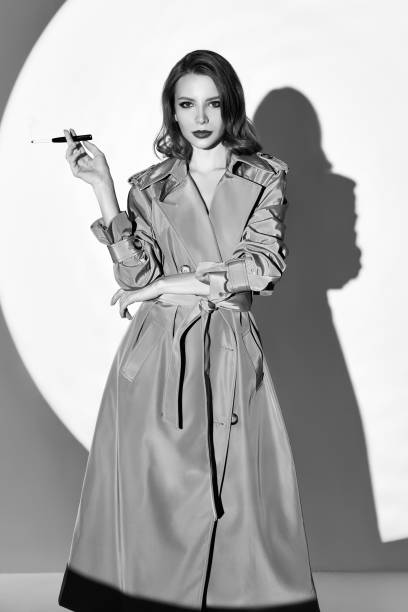Film noir style: beautiful elegant woman smoking cigarette. Vintage (retro) portrait of charming young girl in trench coat. Black and white stock photo