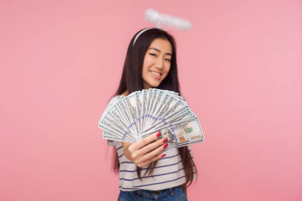 Financial wealth. Portrait of happy smiling girl with halo over head holding out money to camera Financial wealth. Portrait of happy smiling girl with halo over head holding out money to camera, boasting big profit, lottery winning, salary increase. indoor studio shot isolated on pink background bills saints stock pictures, royalty-free photos & images