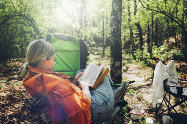 Finding solitude in wilderness concept. Young woman resting in forest, sitting in camp chair and reading book Finding solitude in wilderness concept. Young woman resting in forest, sitting in camp chair and reading book bushcraft stock pictures, royalty-free photos & images