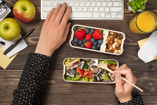 First person top view photo of female hands eating healthy food from two lunchboxes and typing on keyboard mouse glass of juice apples plant and stationery on isolated dark wooden table background