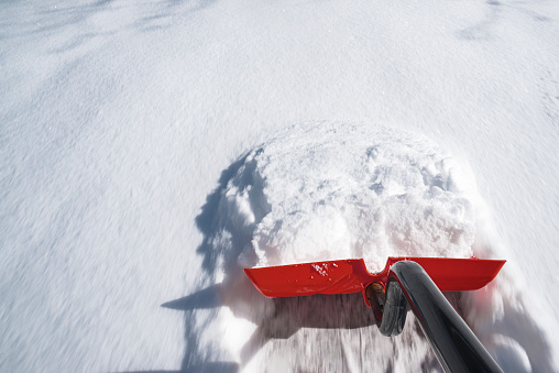 A first-person perspective looking down the handle of a red shovel pushing snow off of a driveway.