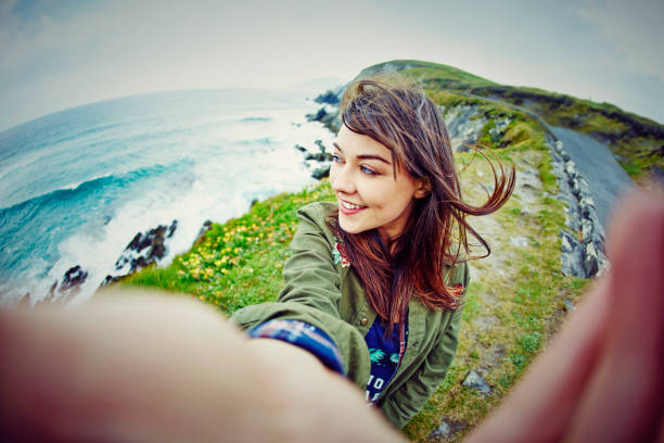 Fish-eye lens of woman taking selfie on mountain by sea Fish-eye lens of thoughtful woman taking selfie while sitting on mountain. Beautiful tourist with windswept brown hair is on grassy cliff by sea. She is wearing casuals during vacation. fish eye lens stock pictures, royalty-free photos & images