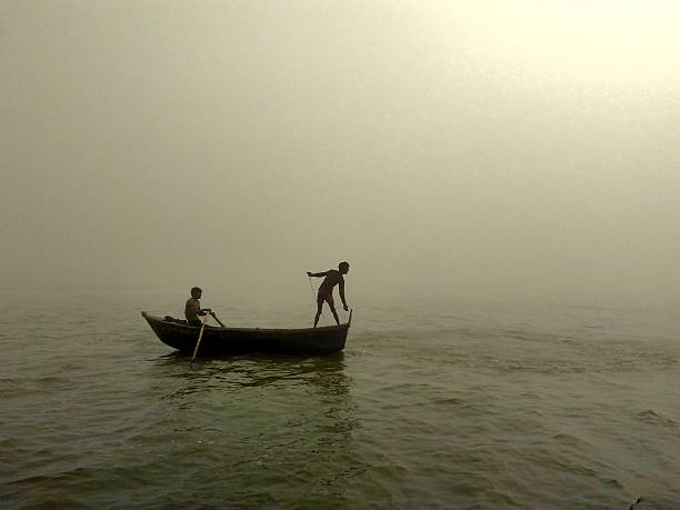 Fishing in winter fog in river Ganges This picture was captured at Ganga Bairaj (Dam over river Ganges) in kanpur, India. It shows a boy throwing fishing net to catch fish in foggy winter morning. And other sitting behind and rowing the boat.  ganges river stock pictures, royalty-free photos & images