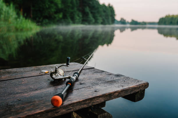 Fishing rod, spinning reel on the background pier river bank. Sunrise. Fog against the backdrop of lake. Misty morning. wild nature. The concept of rural getaway. Article about fishing day. stock photo