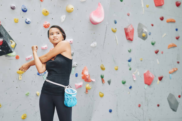 Fit ethnic woman stretching before climbing Young Asian woman in black sportswear and with sack of talk on waist warming up hands before climbing wall in gym bouldering stock pictures, royalty-free photos & images
