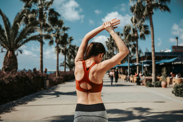 Fit girl doing sport exercise outside on seafront Concentrated slim young woman is raising her arms while warming up before jogging. She is standing while turning her back to camera. Summer activity concept bra stock pictures, royalty-free photos & images