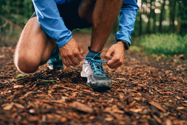 Fit male jogger ties shoes while day training for cross country forest trail race in a nature park stock photo