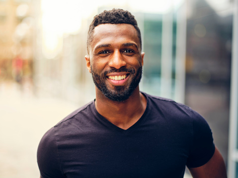 A fit black man with a beard, standing outdoors in a city area.