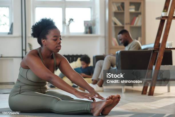 Fit young African woman in tracksuit sitting on the floor of living-room during physical exercise