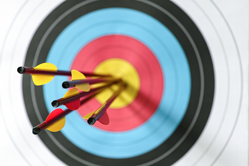 Five arrows in the bull's-eye of an archery target. Selective focus with the focus being on the back end of the arrow, with an out of focus target in the background. Concept image being on target, strategy, aim, accomplishment, aiming for the bull's eye etc.