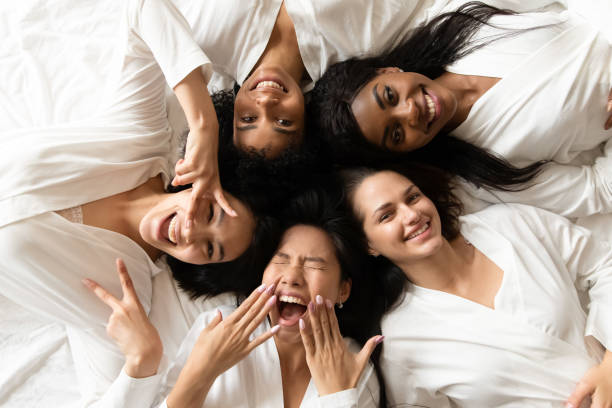 Five happy diverse young girls lying on bed, top view Five happy pretty diverse young girls friends wear robes lying in circle on bed laughing having fun, smiling multi ethnic ladies looking at camera celebrate hen bachelorette party, top view portrait female friendship stock pictures, royalty-free photos & images