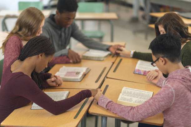 Five students praying Five students in a classroom are sitting with their desks pushed together. They are holding hands in a circle and have their heads bowed in prayer. There are bibles open on their desks. catholicism stock pictures, royalty-free photos & images