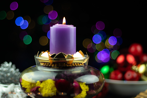 Flames Lit From Designer Purple Or Lilac Pillar Candle Placed Glass Bowl Filled With Flowers And Colorful Bokeh On Dark Black Background. Theme For Shubh Deepawali, Merry Christmas And Happy New Year