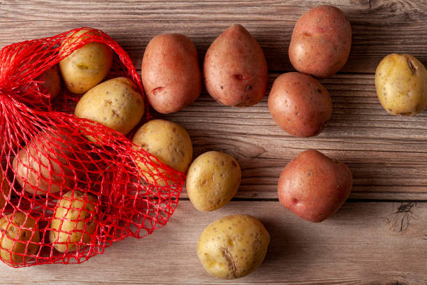 Flat lay  close up image featuring a red mesh potato sack with pink and yellow raw organic potatoes on wooden background stock photo
