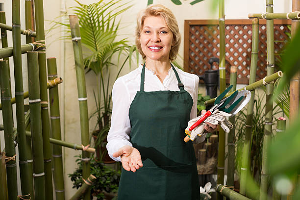 Florist with tools in floral shop Portrait of friendly smiling mature woman florist with tools in hands among green plants in floral shop  How to set up your gardening service stock pictures, royalty-free photos & images