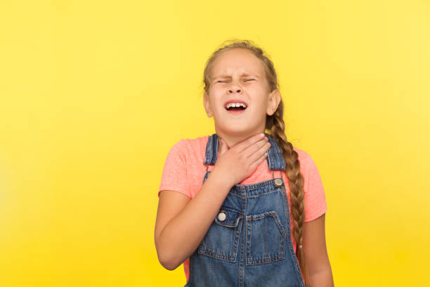 Flu symptoms, neck pain. Portrait of sick little girl in denim overalls suffering sore throat or tonsillitis, thyroid disorder Flu symptoms, neck pain. Portrait of sick little girl in denim overalls suffering sore throat or tonsillitis, thyroid disorder, medical problem. indoor studio shot isolated on yellow background choking photos stock pictures, royalty-free photos & images