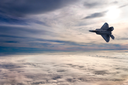 An F22 fighter jet soars above the clouds as the sun begins to set over a beautiful cloudscape.