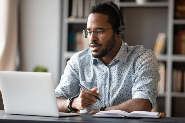 de geconcentreerde afrikaanse zakenman draagt hoofdtelefoonsstudie online het letten op webinar - laptop computeren stockfoto's en -beelden