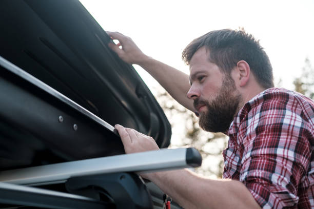 focused bearded man peers into an open roof rack on a car or in a cargo box. preparing to leave. - fechar porta bagagens imagens e fotografias de stock