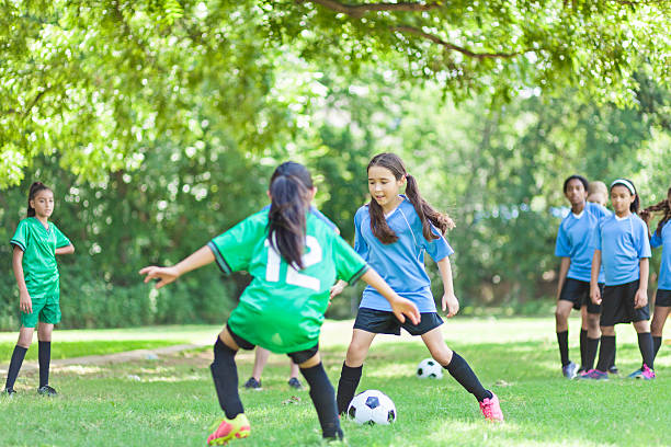 Focused female  soccer players face off Intense young female soccer players are competing. The player in the green jersey blocks the ball as the player in the blue jersey prepares to kick the ball. Opposing team members are in the background. Trees line the playing field. defending sport stock pictures, royalty-free photos & images