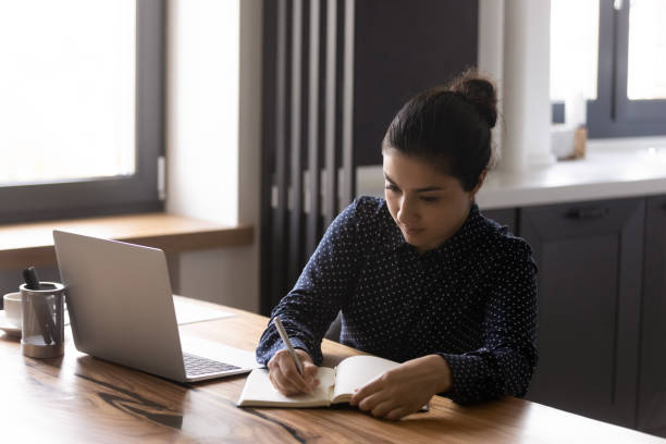 Focused Indian woman make notes studying online on laptop