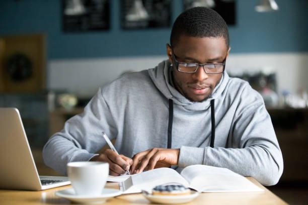 Focused millennial african student making notes while studying in cafe Focused millennial african american student in glasses making notes writing down information from book in cafe preparing for test or exam, young serious black man studying or working in coffee house literature stock pictures, royalty-free photos & images