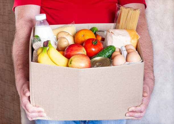 Food box in male hands Grocery delivery courier man in red uniform holds cardboard box with fresh vegetables, fruits and other food. Express food delivery, donation concept. food bank stock pictures, royalty-free photos & images