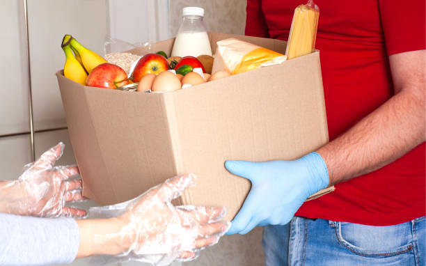 Food delivery during quarantine Grocery delivery courier man in red uniform and medical gloves gives box of food to girl in protective gloves near door. Safe food delivery during quarantine, online shopping or donation concept. food donation stock pictures, royalty-free photos & images