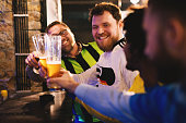 Football fans in jerseys of their teams are saluting to each other with beer while sitting at the bar.