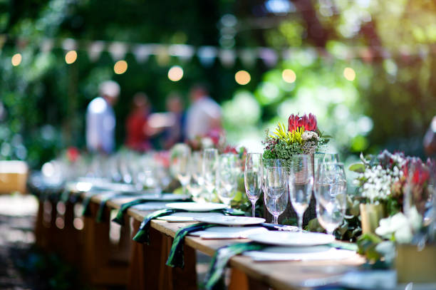 Forest Table Setting with flowers A protea on an outdoor wooden table set for a celebration in a forest during daytime with plates, glasses, napkins, fynbos flower arrangements and string lights and bunting defocussed with people behind Cape Town South Africa banquet photos stock pictures, royalty-free photos & images