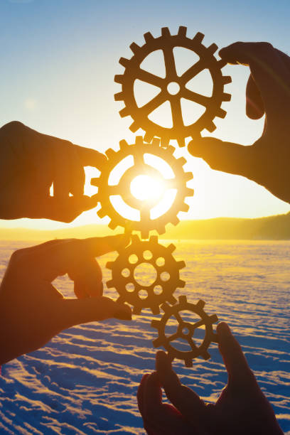 Four hands of businessmen holding gears folded into a puzzle mechanism at sunset. stock photo