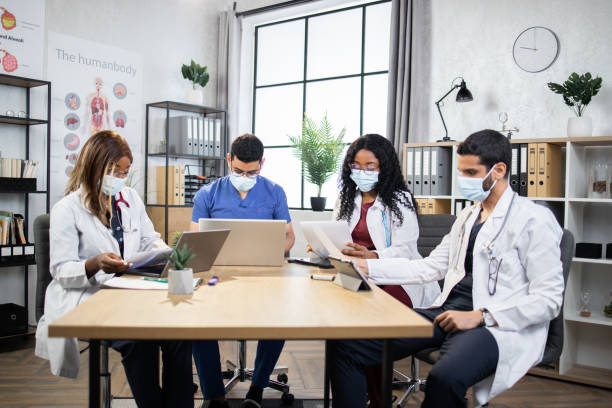 Four multiracial doctors examining medical record of patient, working in modern office Group of four multiracial doctors in masks using modern gadgets while examining patient's record during consilium. International colleagues sitting at room discussing various way of treatment. medical student stock pictures, royalty-free photos & images