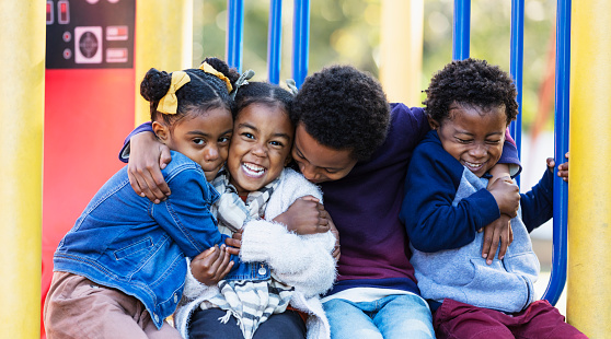 Four siblings sitting at the playground, hugging and smiling at the camera. The oldest is an 8 year old boy. The girl on the left is 5 years old, and the youngest children are 3 year old twins. They are mixed race Hispanic, African-American and Native American.