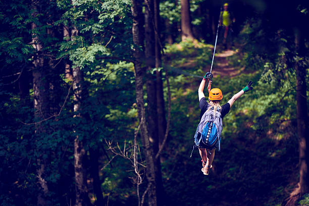 freedom and adventure rear view of young woman in the forest enjoying the adventure on the zip line, having fun and holding her arm raised. adrenaline stock pictures, royalty-free photos & images
